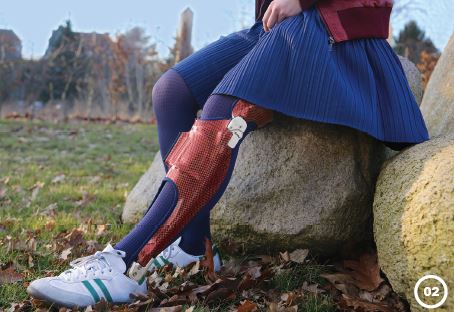 Frau mit blauem Rock und blaue Strümpfe trägt rote Orthese. Sie sitzt auf einem Stein