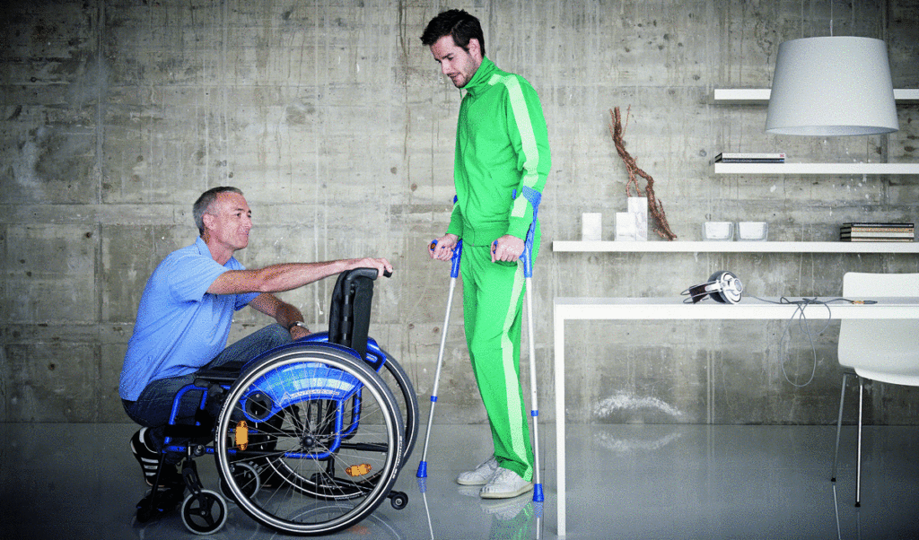 Rehatechniker präsentiert Rollstuhl einem jungen Mann mit Unterarmgehstützen